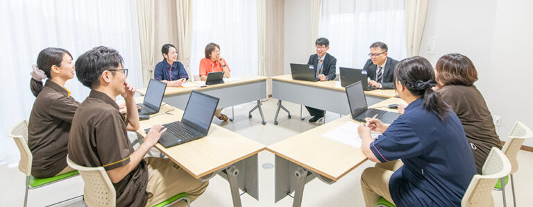 介護施設のスタッフによる会議・研修風景。複数の職員がノートパソコンを前にした会議テーブルに着席している。