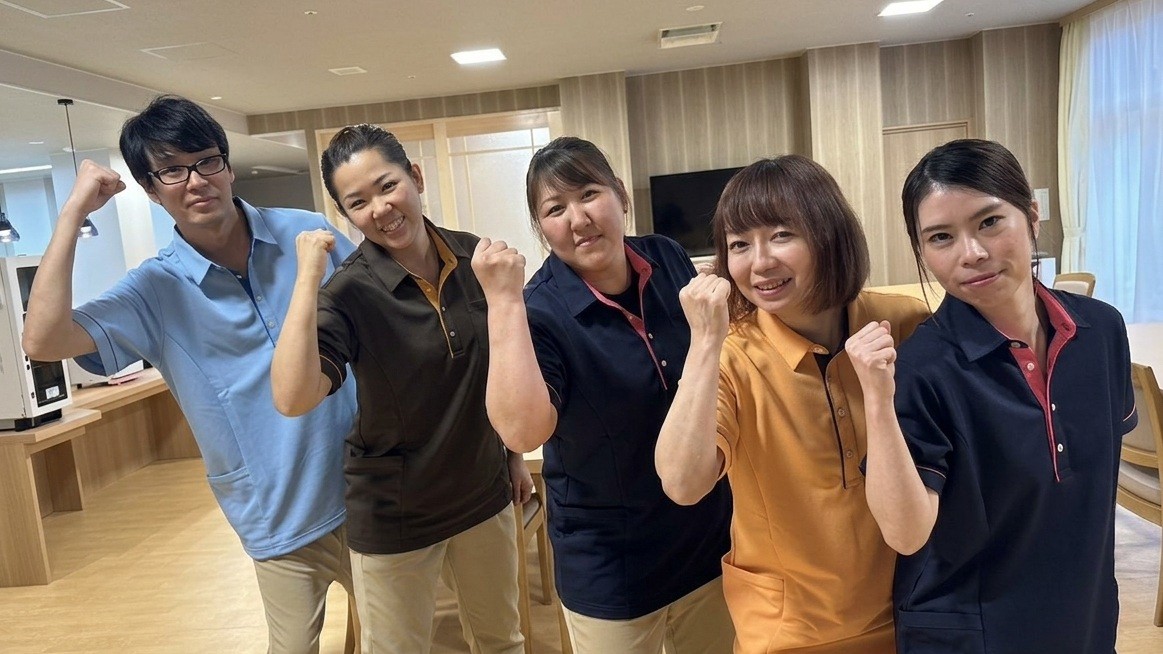 介護施設のスタッフ5名が笑顔で一堂に集まり、ガッツポーズをしている風景